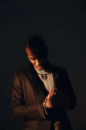 A man in a suit and sweater poses in a studio with dark lighting, showcasing fashion and style with a contemplative expression.