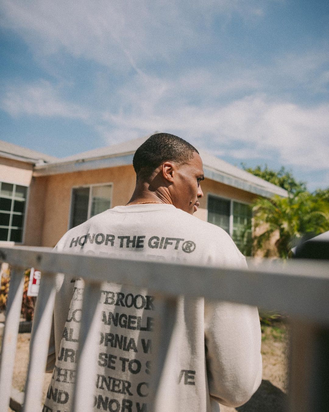 An adult male is seen from the back in a cream-colored sweater with black text standing outside a peach stucco house under a partly cloudy sky.