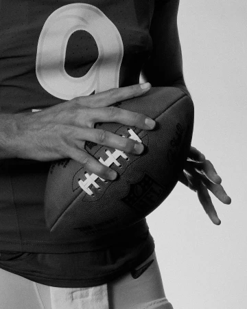 Close-up shot of an adult male wearing a jersey with the number nine, holding an American football with both hands in a studio setting.