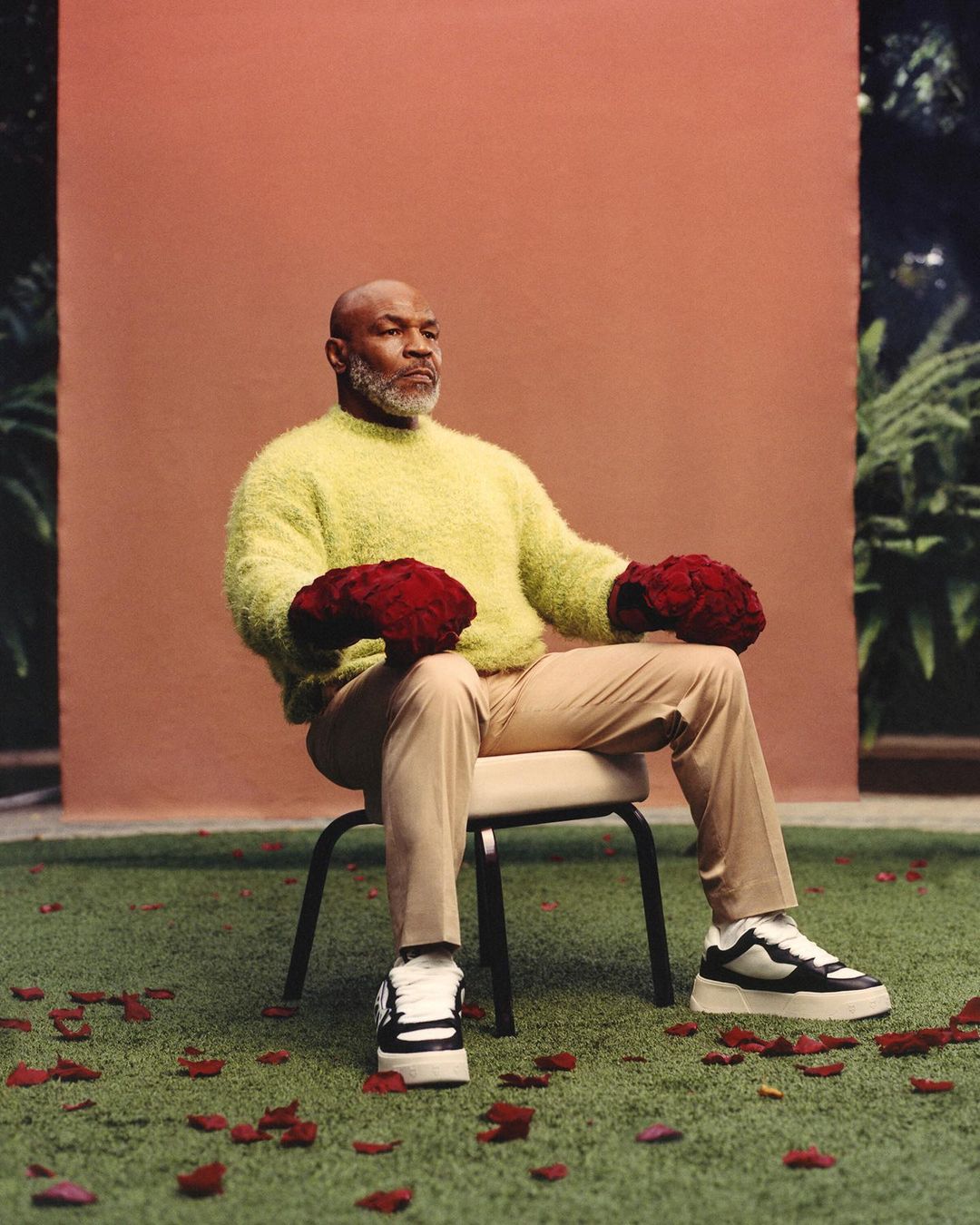 Mike Tyson sits in a chair on a lawn with rose petals, holding red rose boxing glove bouquets, wearing a fuzzy yellow sweater and sneakers.