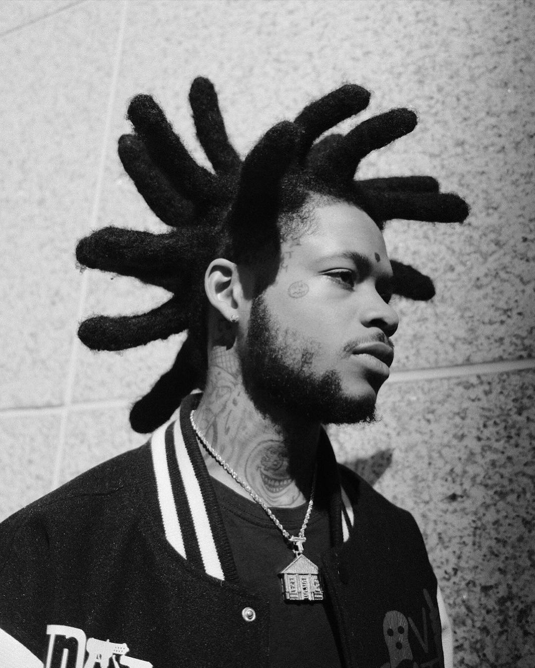 Black and white portrait of a black male with unique hairstyle, facial tattoo, necklace, and bomber jacket against a neutral wall.
