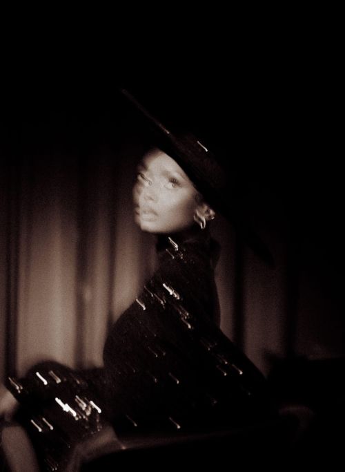 Portrait of a stylish African American woman in a black hat and turtleneck dress with light bokeh effect in a dark, moody ambiance.