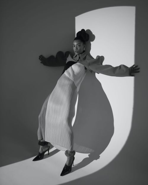 Black and white portrait of a woman in a sculptural pleated dress posing in a light-wash studio with a circular light effect.
