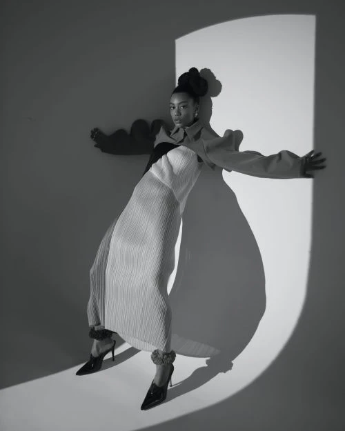 Black and white portrait of a woman in a sculptural pleated dress posing in a light-wash studio with a circular light effect.