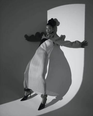 Black and white portrait of a woman in a sculptural pleated dress posing in a light-wash studio with a circular light effect.