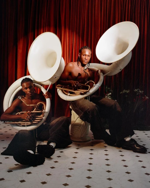 Two shirtless African-descent men with sousaphones in a studio with a red curtain and pattern floor, wearing pants and shoes.