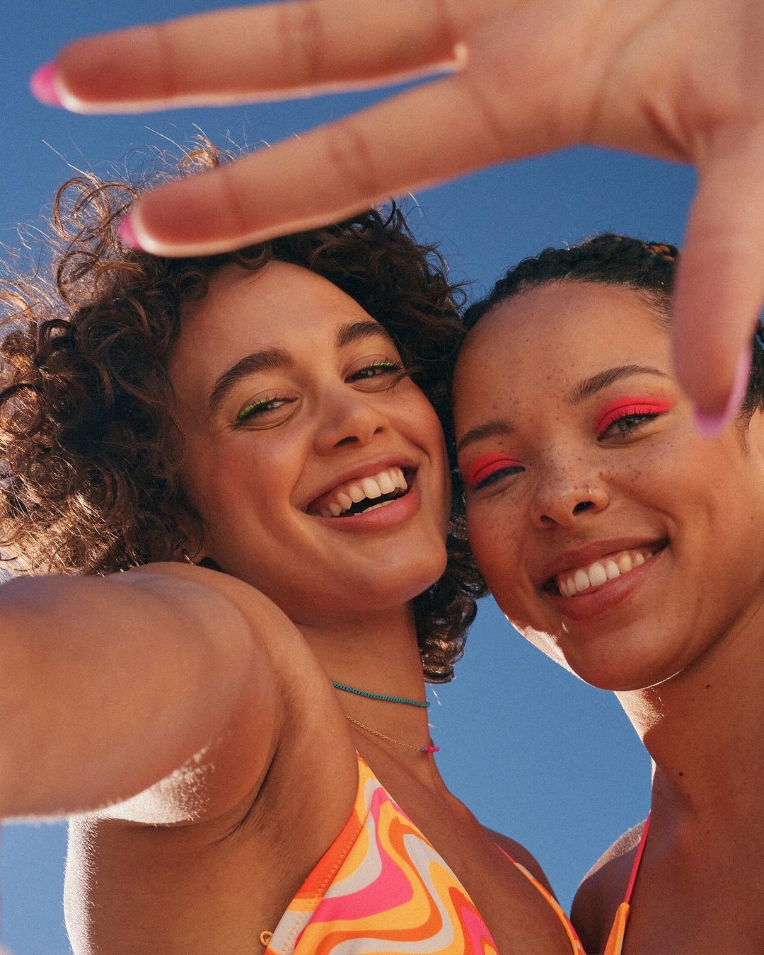Two smiling young women wearing colourful swimsuits are posing outdoors against a blue sky, framing their faces with a hand gesture.
