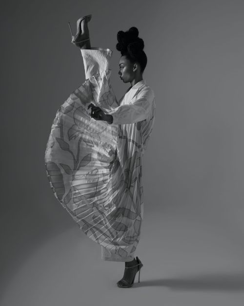 Monochrome portrait of a Black woman posing in a studio, wearing patterned dress and high heels with one leg extended vertically.
