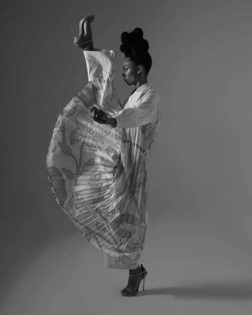 Monochrome portrait of a Black woman posing in a studio, wearing patterned dress and high heels with one leg extended vertically.