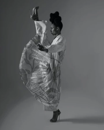 Monochrome portrait of a Black woman posing in a studio, wearing patterned dress and high heels with one leg extended vertically.