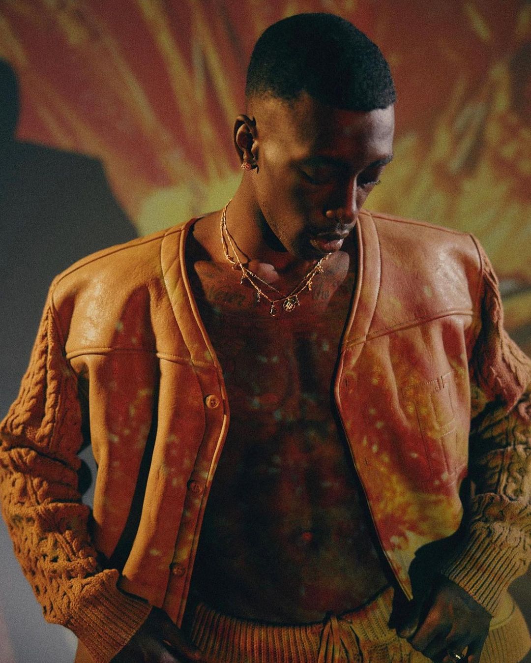 Man in open tan leather jacket, knit sleeves, wearing jewelry, looks down against background with red and yellow abstract pattern.