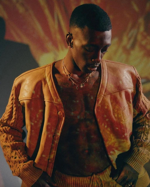 Man in open tan leather jacket, knit sleeves, wearing jewelry, looks down against background with red and yellow abstract pattern.