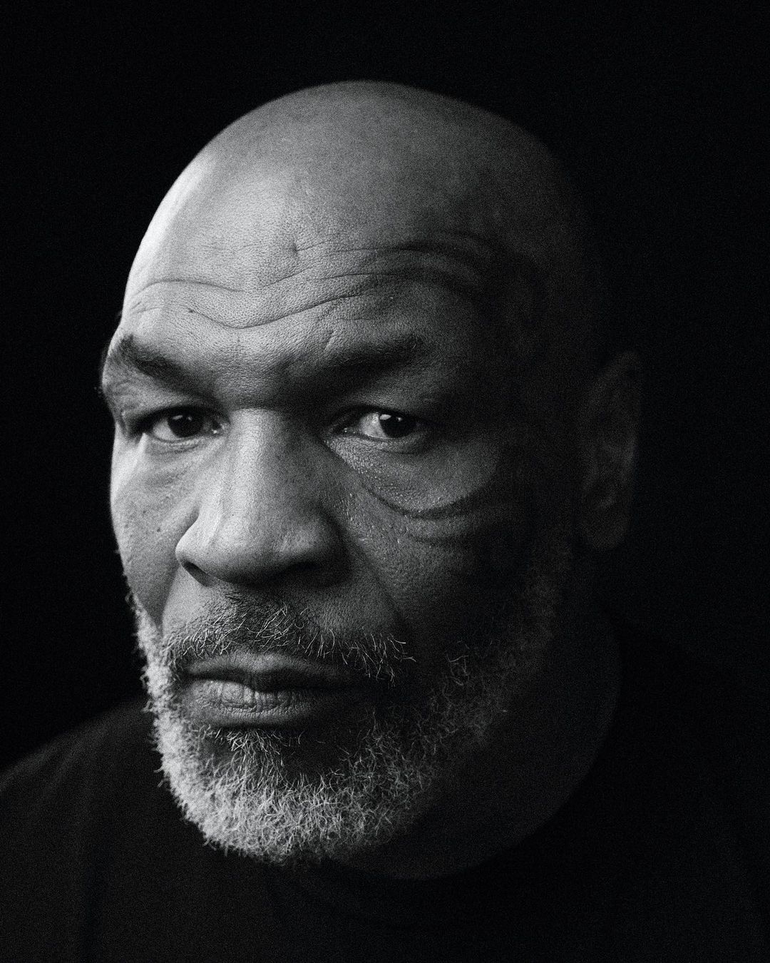 Black and white portrait of adult male with bald head and distinctive face tattoo on black background looking towards camera.