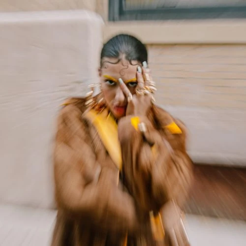 Woman in a brown fur coat with bold yellow makeup and statement jewelry posing in front of a neutral brick wall with a window.