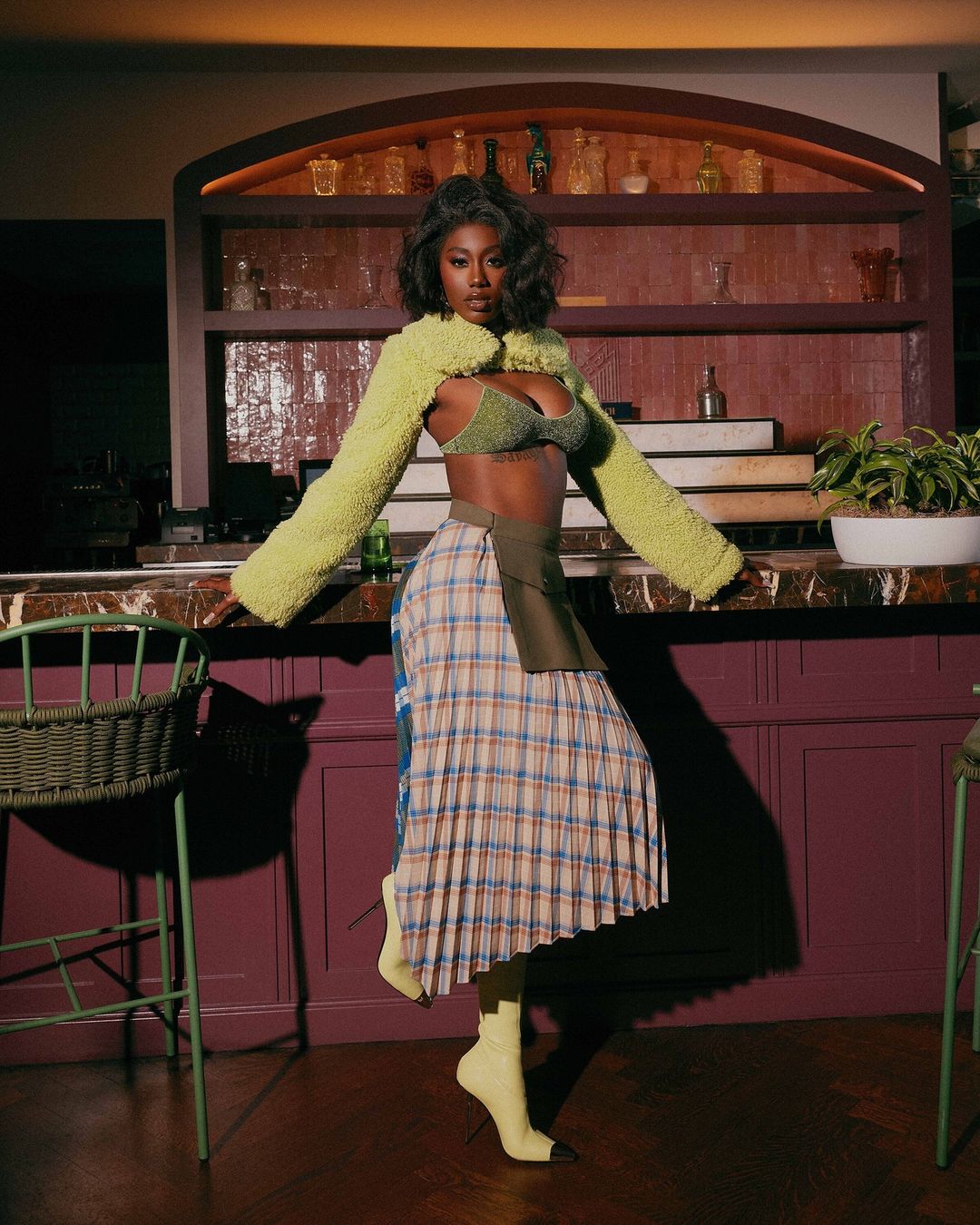 Adult female poses indoors in a plaid skirt, green bra top and bolero. Behind is a bar area with wine bottles and plants.