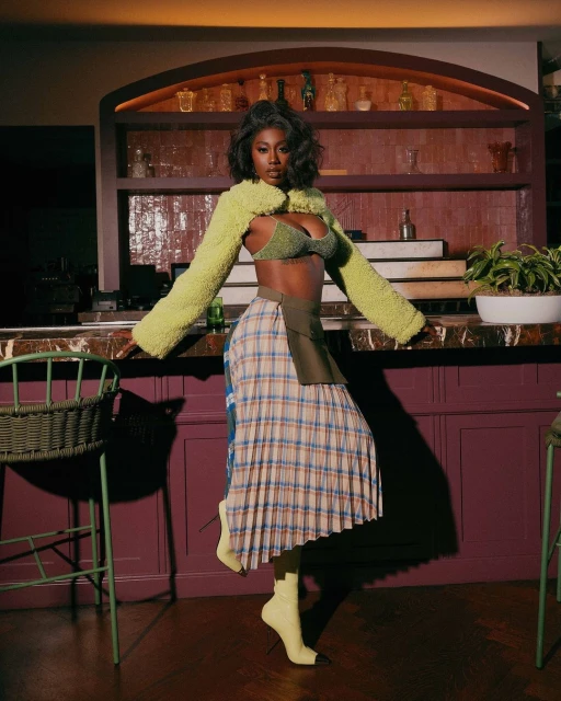Adult female poses indoors in a plaid skirt, green bra top and bolero. Behind is a bar area with wine bottles and plants.