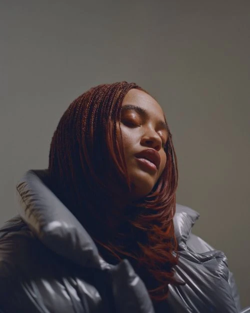 Portrait of an adult woman with eyes closed, styled hair, wearing a puffy jacket, and set against a plain backdrop in a studio setting.