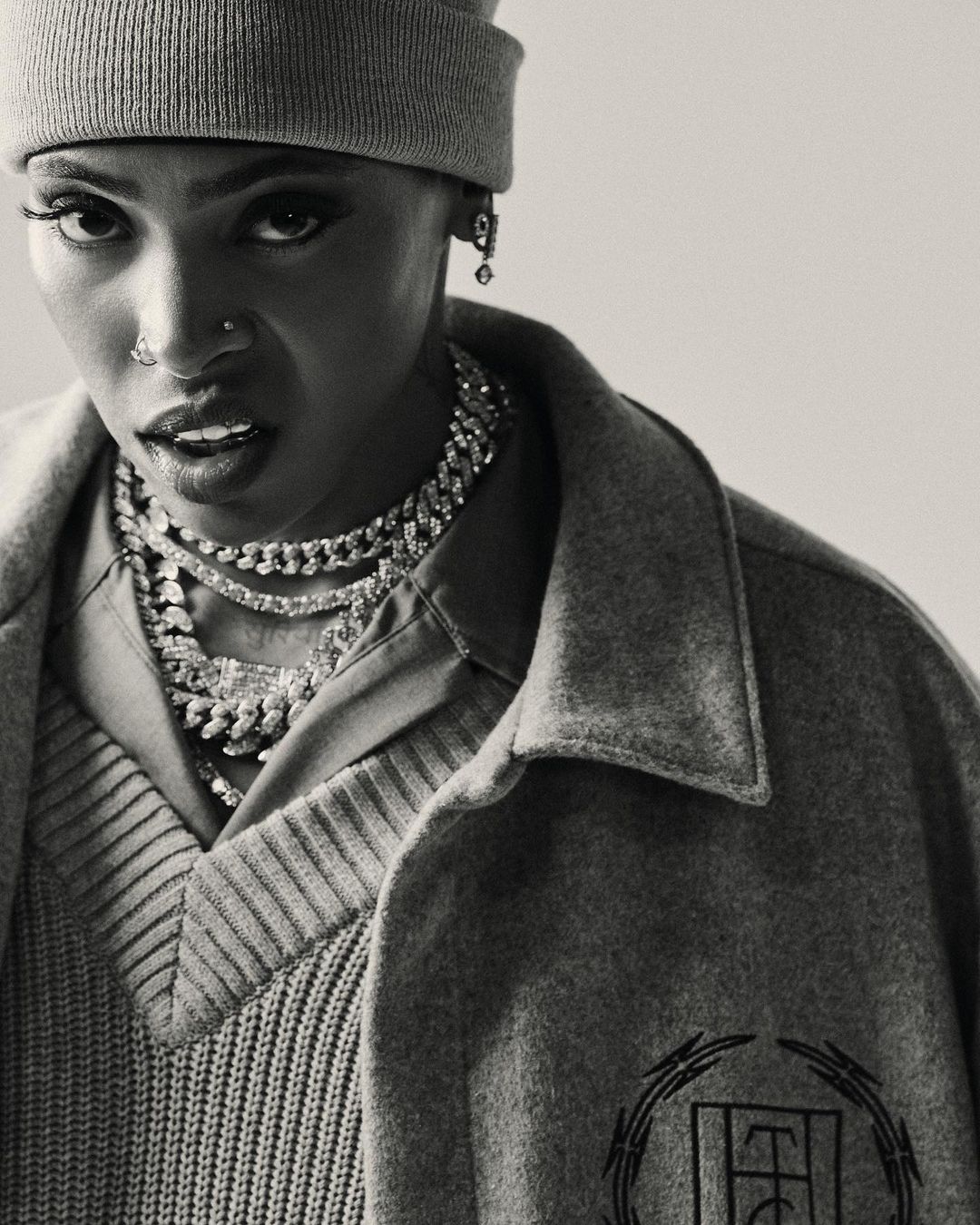 Portrait of a short-haired person wearing a grey beanie, statement necklaces, and grey outerwear in a studio with soft lighting.