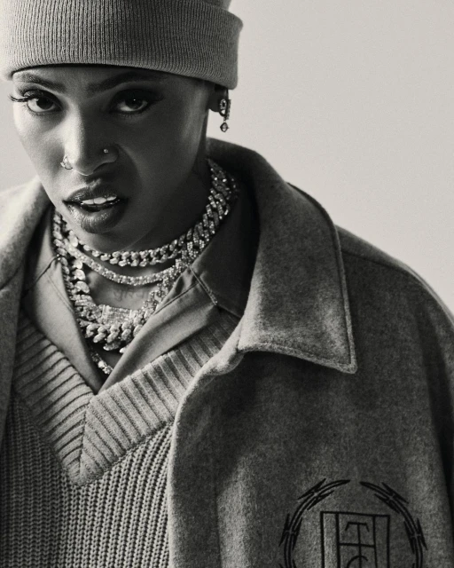 Portrait of a short-haired person wearing a grey beanie, statement necklaces, and grey outerwear in a studio with soft lighting.