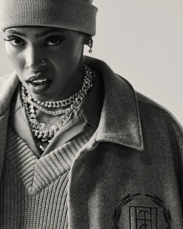 Portrait of a short-haired person wearing a grey beanie, statement necklaces, and grey outerwear in a studio with soft lighting.