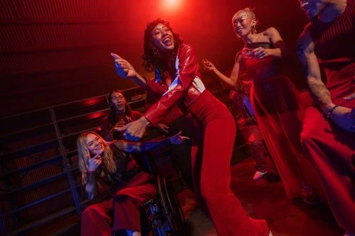 Group of diverse people dancing in red-lit venue; woman in wheelchair; fun, energetic vibe, vivid red hues, club setting, event photography.