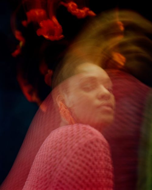 Portrait of a person with eyes closed, wearing a pink sweater, and featuring blurred motion and dark background with a red pattern.