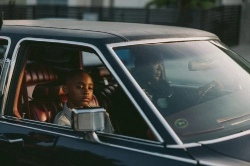 A child sits in the passenger seat of a car, with an adult driving. The vintage car is dark colored and reflects outdoor light.