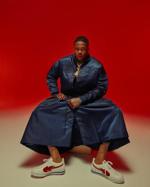 Portrait of a seated adult male wearing a blue satin jacket and skirt, with white sneakers against a red backdrop. 