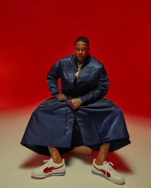Portrait of a seated adult male wearing a blue satin jacket and skirt, with white sneakers against a red backdrop. 