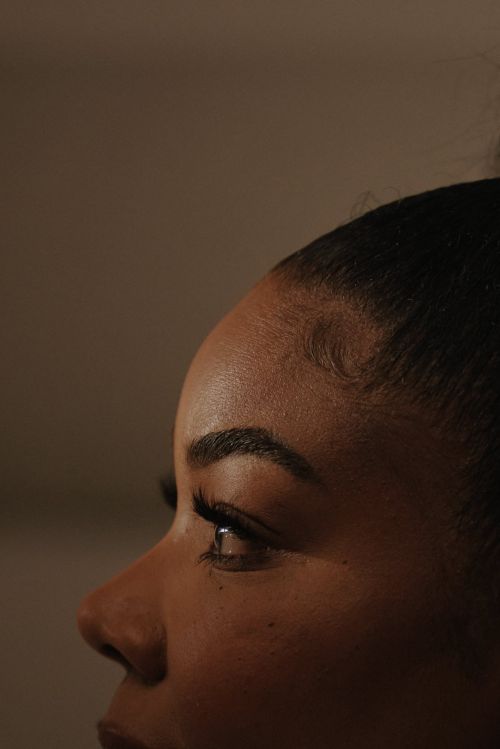Close-up profile portrait of a Black woman with styled hair edges and dark eyelashes against a dark background
