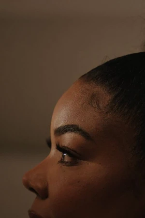 Close-up profile portrait of a Black woman with styled hair edges and dark eyelashes against a dark background