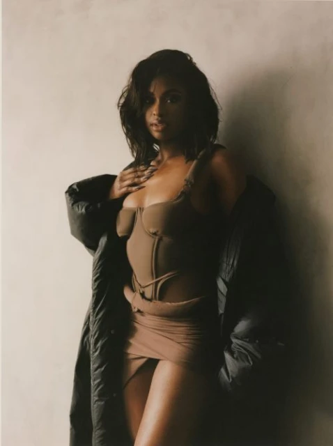 Attractive woman with brown hair poses in a corset top and mini skirt, wearing an oversized jacket against a neutral backdrop.