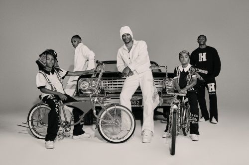 Group of five young men in black and white outfits, posing with lowrider bicycles in front of a vintage car in a studio setting.