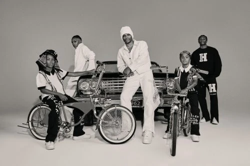 Group of five young men in black and white outfits, posing with lowrider bicycles in front of a vintage car in a studio setting.