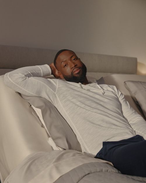 Adult male with beard reclining on a bed with a white shirt and blue pants in a casual setting, looking at the camera.