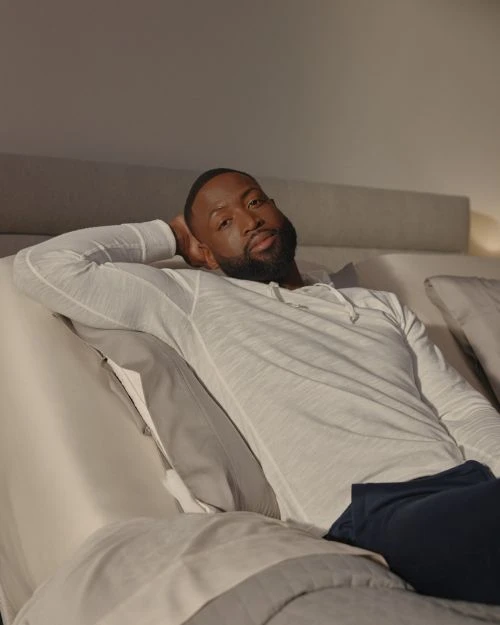 Adult male with beard reclining on a bed with a white shirt and blue pants in a casual setting, looking at the camera.