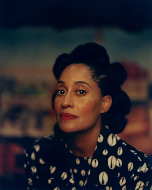 Close-up portrait of an adult woman with dark hair styled up, wearing a patterned top against a blurred painted backdrop.