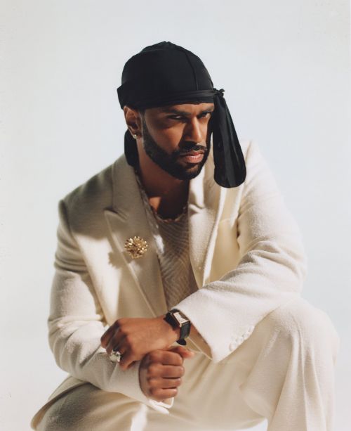 Portrait of stylish adult male in a white suit and black durag in a photo studio. He is wearing jewelry and a watch.