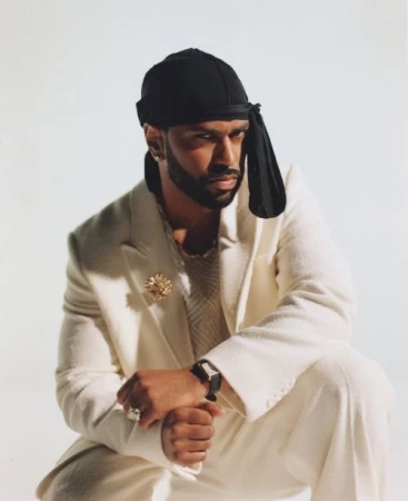 Portrait of stylish adult male in a white suit and black durag in a photo studio. He is wearing jewelry and a watch.