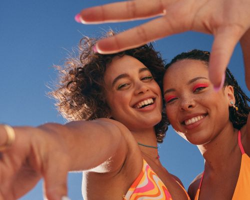 Two smiling women pose outdoors against a blue sky, showcasing vibrant makeup and summer fashion, with one woman framing the shot with her hand.