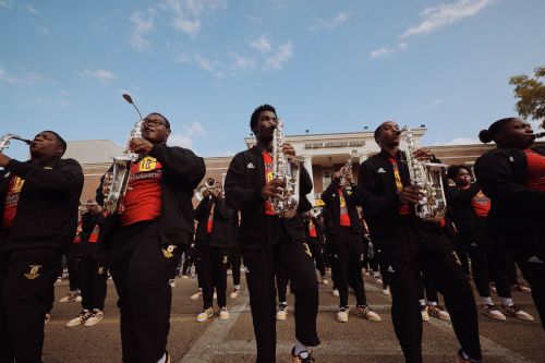 Marching band performing outdoors on street with saxophones and black and gold uniforms under a bright blue sky. 