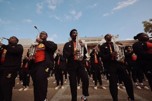 Marching band performing outdoors on street with saxophones and black and gold uniforms under a bright blue sky. 