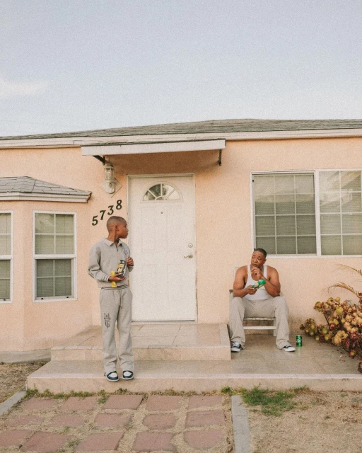 Two adult males pose outside a peach-colored house with white trim, one standing on the stairs and the other sitting on the porch.