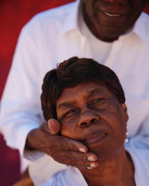 Portrait of a Black woman with a warm smile, being embraced by an older Black man in a white shirt against a vivid red background.