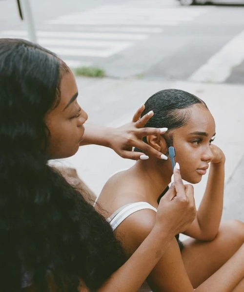 Two young African-American women styling hair outdoors on an overcast day, one applying a grooming tool, showcasing beauty and personal care.