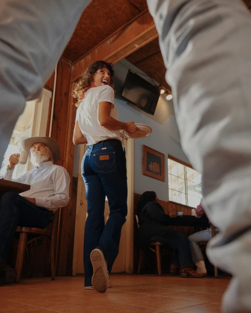 An indoor diner scene with a smiling woman in a white t-shirt, blue jeans, and carrying a plate. Seated customers and wooden interiors.
