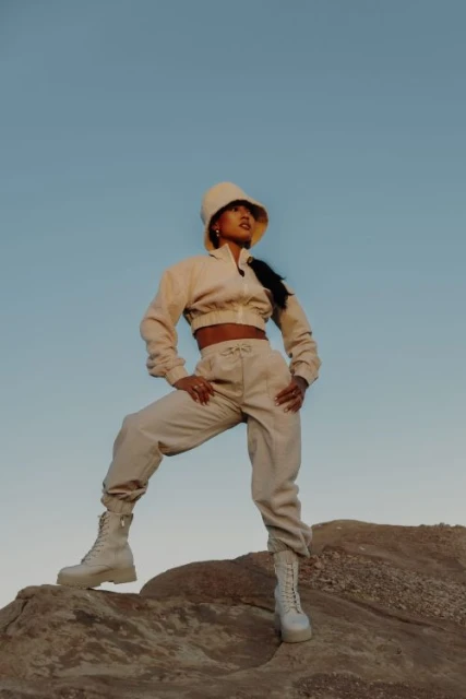 Woman poses in a sandy outdoor landscape wearing a bucket hat, cropped jacket, pants, and boots against a light blue sky backdrop.