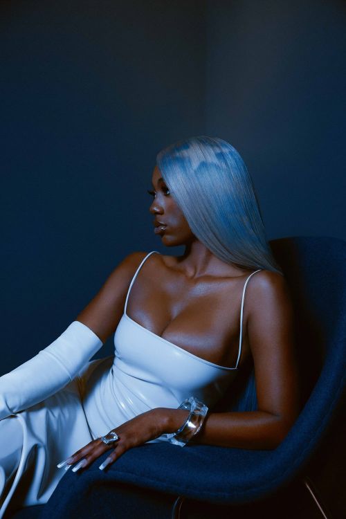 Glamorous woman with silver hair poses in white dress with arm-length gloves, seated in a blue chair against a dark blue background.
