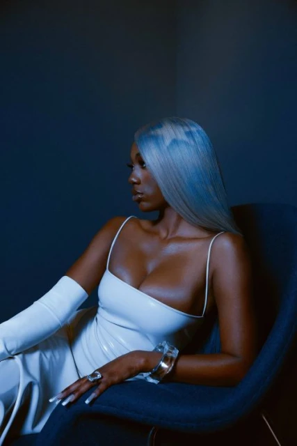 Glamorous woman with silver hair poses in white dress with arm-length gloves, seated in a blue chair against a dark blue background.