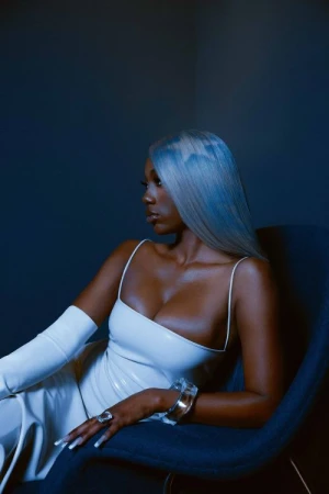 Glamorous woman with silver hair poses in white dress with arm-length gloves, seated in a blue chair against a dark blue background.